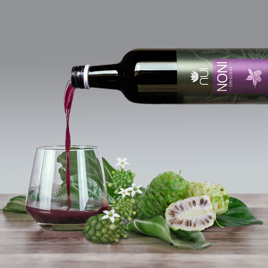 NUI NONI Original Saft wird aus einer Flasche in ein Glas eingeschenkt, auf dem Tisch liegen Noni Früchte