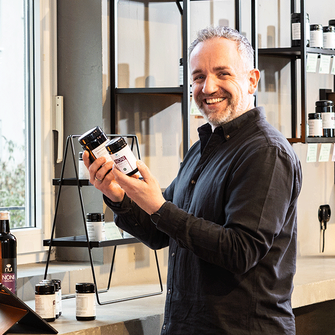 „Mitarbeiter hält zwei NUI Noni Produktdosen in der Hand und steht in einem Raum mit Regalen.“